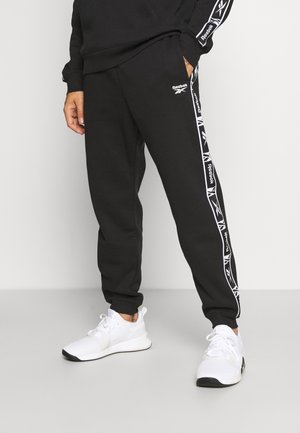 Osoba nosící černé tepláky Reebok s bílými proužky s logem po stranách a bílé sportovní boty na šedé podlaze.