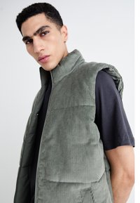 Vesta verde din corduroy cu guler înalt, secțiuni quilted și un fermoar frontal, purtată peste o cămașă neagră. Textura prezintă dungi verticale.