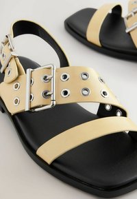 Sandale plate negre și galbene cu o curea lată pentru degete, cataramă argintie și detalii cu eyelet-uri. Partea superioară are o textură de piele netedă și o talpă plată.
