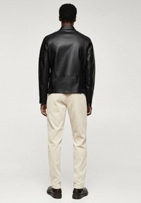 Giacca di pelle nera con un design aderente, texture liscia e colletto alto. Abbinata a pantaloni avorio e scarpe marroni.