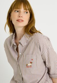 Camisa de rayas marrón y blanca con cuello abotonado, mangas cortas y un diseño bordado de un perro en el bolsillo.