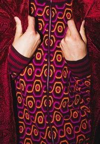 Pull à motifs colorés avec fermeture éclair : formes géométriques audacieuses en violet, orange et noir ; poignets côtelés ; porté sous une couche extérieure bordeaux riche.