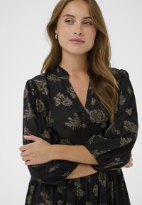 Rochie neagră cu modele florale în tonuri estompate, având decolteu în V și mâneci bufante. Realizată dintr-un material moale, cu detalii pliate.
