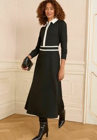 Vestido negro de manga larga con cuello blanco y acentos en la cintura, tejido suave, diseño ajustado y acampanado, combinado con botas de tacón negras hasta la rodilla.
