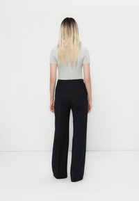 Persoană cu părul lung blond stă cu fața spre un perete alb, purtând un top scurt ajustat gri și pantaloni negri cu picioare largi.
