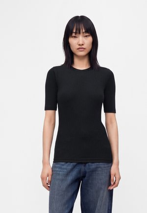 Kvinde med skulderlangt sort hår iført en sort ribbet kortærmet top og blå jeans, stående foran en ensfarvet hvid baggrund.