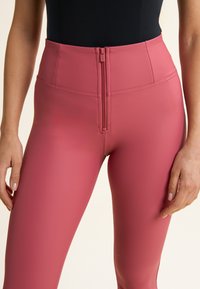 Leggings rosas de tiro alto, textura suave, acabado mate, con cremallera frontal, ajuste entallado y detalles cosidos a lo largo de las costuras.