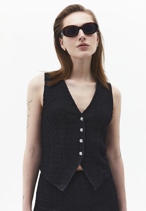 Jeune femme portant des lunettes de soleil ovales à monture écaille, un gilet sans manches noir texturé à boutons, et de petites boucles d'oreilles créoles dorées.