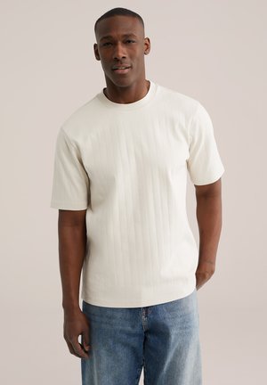 Homme debout portant un t-shirt côtelé à manches courtes de couleur blanc cassé uni et un jean bleu, sur un fond neutre.