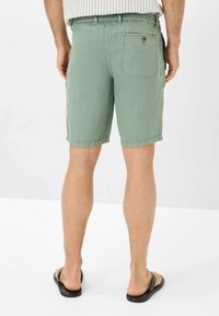 Pantaloni corti verdi in cotone con cintura elastica e tasca posteriore. Orlo che arriva a metà coscia. Indossati con infradito nere.
