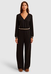 Schwarzer Jumpsuit mit langen Ärmeln, V-Ausschnitt, elastischem Bund in der Taille und einem kontrastierenden beigen Taillenbund. Weicher Stoff, weit geschnittene Beine und vielseitiger Stil.