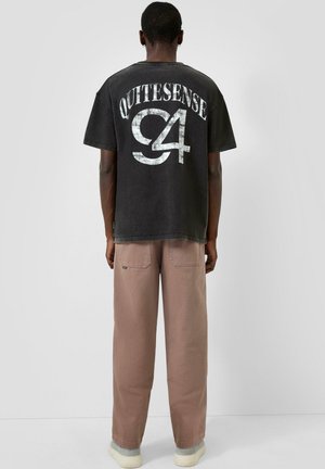 Person wearing black oversized t-shirt with white "QUITESENSE C4" text on back, paired with loose brown pants and light-colored shoes.