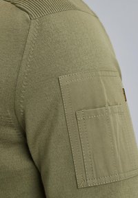 Olivno zeleni pulover z teksturirano pleteno telo, rebrastim ovratnikom ter kvadratno prsno žepom z vidnimi šivi.