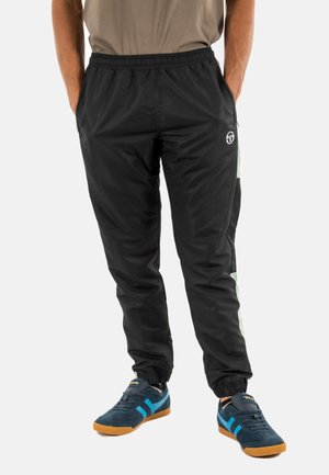 Hombre vistiendo pantalones de chándal negros con rayas blancas en los lados y zapatillas de deporte azul marino con detalles azules, de pie con las manos en los bolsillos.