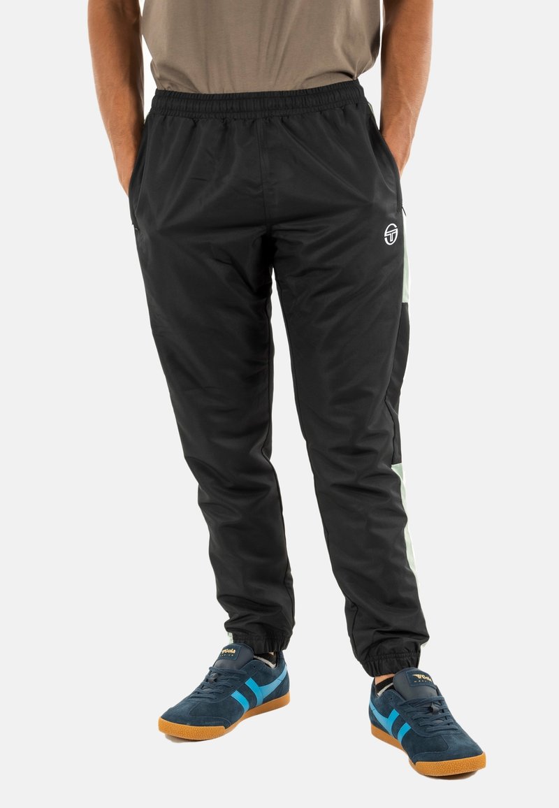 Man in zwarte trainingsbroek met witte zijkanten en donkerblauwe sneakers met blauwe accenten, staande met handen in de zakken.