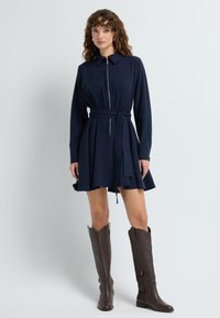 Granatowa sukienka z kołnierzykiem i zamkiem z przodu, wcięciem w talii i rozkloszowaną spódnicą. Połączona z brązowymi kozaczkami do kolan. Gładka tekstura materiału.