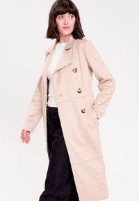 Cappotto beige in tessuto morbido, con un design doppiopetto, grandi bottoni e tasche laterali. Indossato sopra una maglietta bianca e jeans scuri.