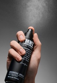 Bottiglia aerosol nera con finitura opaca, ugello rotondo, tenuta da una mano, diretta verso l'alto, spruzzo di nebbia visibile su uno sfondo grigio.