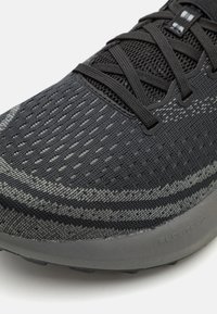 Zapatilla deportiva negra con parte superior de punto texturizado, que presenta franjas horizontales grises y cordones negros. Incluye un collar acolchado y una suela de goma.