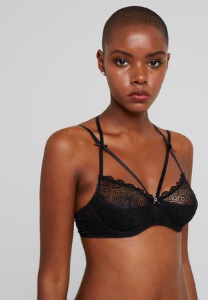 Zwarte kant bralette met een geometrisch patroon, golvende randen en dunne verstelbare bandjes. Centraal strikdetail met een kleine decoratieve charme.