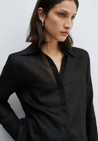 Sort gennemsigtig bluse med krave, lange ærmer og en V-formet front. Teksturen er let og let gennemsigtig.