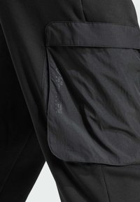 Black jogger pants featuring a large, textured side pocket in a contrasting matte finish, with printed branding and a sleek, streamlined design.