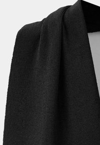 Écharpe noire en tricot suspendue avec une texture côtelée visible et des plis de tissu doux sur un fond clair.