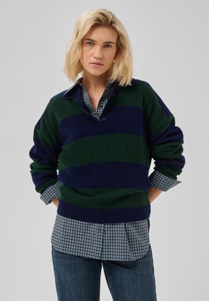 Femme blonde portant un pull rayé vert foncé et bleu marine par-dessus une chemise à carreaux et un jean bleu, debout les mains dans les poches.