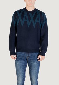 Homme portant un pull bleu marine avec des motifs en zigzag teal sur la poitrine et les épaules, associé à un jean bleu délavé.