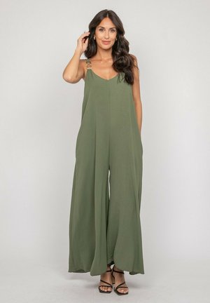 Femme aux longs cheveux foncés portant une combinaison pantalon kaki à jambes larges avec des détails en anneau sur les bretelles et des sandales à talons noires, debout devant un fond gris.