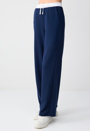 Person trägt marineblaue, weit geschnittene Hose mit weißem elastischem Bund und weißen Turnschuhen, steht vor einem schlichten hellen Hintergrund.