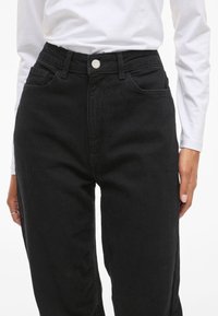 Crne traperice visokog struka s klasičnim dizajnom s pet džepova, šivanim detaljima i srebrnim kopčanjem na struku.