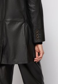 Mustahi leather jacket, joka on varustettu sileällä pinnalla, räätälöidyllä istuvuudella ja nappikiinnityksellä, jossa on neljä mustaa nappia ja metallinen reuna.