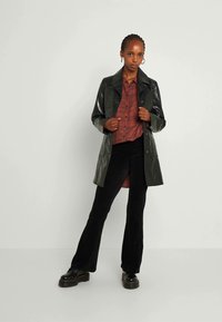Donkergroene, glanzende, knielange trenchcoat over een rood geruit overhemd. Zwarte uitlopende broek en chunky zwarte schoenen maken de outfit compleet.