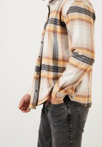 Camisa de franela con botones en patrón a cuadros de color crema, naranja, amarillo y negro. Hecha de tela texturizada, con mangas largas y corte recto.