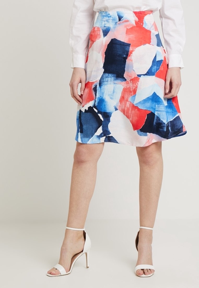 Femme portant une jupe imprimée abstraite mi-longue bleue, rouge et blanche, associée à des sandales blanches à talons hauts avec bride à la cheville.