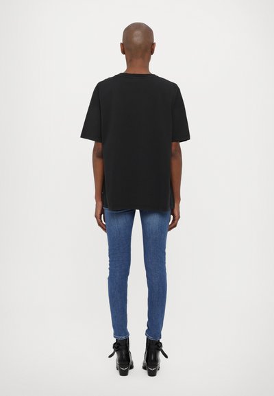 Camiseta negra de manga corta con dobladillo recto, combinada con jeans ajustados azules y botas negras de tobillo, vista desde atrás contra un fondo liso.