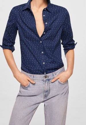 Kvinde iført en marineblå mønstret skjorte med knapper og opkrogede ærmer samt højtsiddende lysegrå jeans, med hænderne i lommerne.