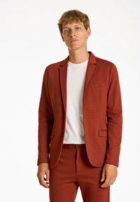 Blazer color ruggine realizzato in un tessuto texturizzato, con rever a tracolla, due tasche frontali e due bottoni, abbinato a una t-shirt bianca.