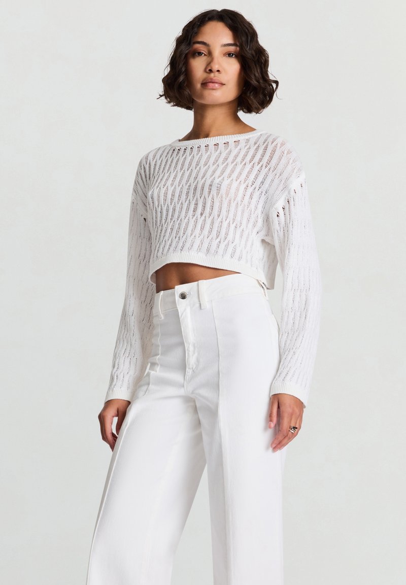 Maglione corto bianco a maglia con motivo a trama aperta, maniche lunghe e polsini a costine, abbinato a jeans bianchi a vita alta e gamba larga.