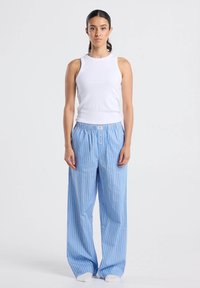 Nuori nainen seisoo valkoisessa hihattomassa paidassa ja rennoissa sinivalkoraidoissa olevissa housuissa, katsoen suoraan eteenpäin vaaleaa taustaa vasten.