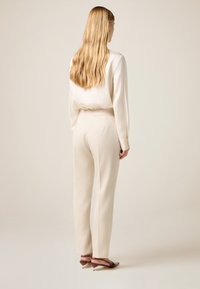 Satin-creme Bluse mit langen Ärmeln, kombiniert mit maßgeschneiderten cremefarbenen Hosen mit elastischem Bund und leichter Textur. Braune Absatzschuhe.