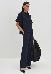 Camicia blu navy con maniche corte, abbinata a pantaloni scuri e una borsa a tracolla nera intrecciata. Tessuto liscio con un motivo testurizzato.