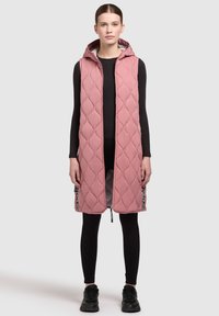 Rosa gesteppte ärmellose Weste mit Kapuze, mit einem Reißverschluss vorne und schwarzen Textakzenten an den Seiten, getragen über schwarzen Leggings.