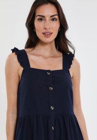 Abito blu navy con spalline ruffle regolabili, scollo quadrato e chiusura con bottoni sul davanti. Presenta una vestibilità ampia e una gonna arricciata.
