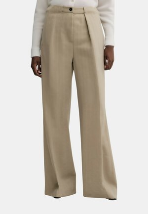 Pantaloni beige a gamba larga e vita alta con chiusura con bottoni, indossati con una maglia bianca a maniche lunghe, mani rilassate ai lati.