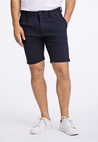 Mörkblå shorts i mjukt tyg med knappstängning, sidfickor och uppvikta fållar, kombinerade med vita sneakers.