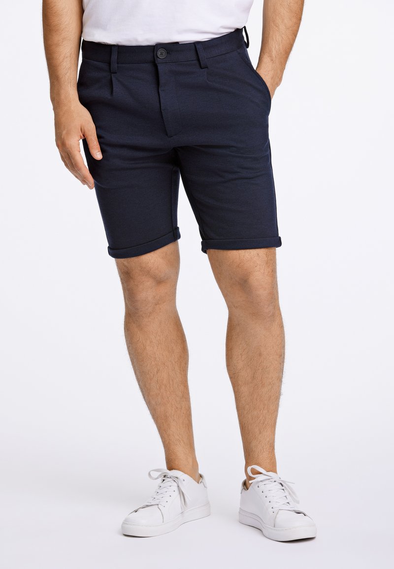 Mörkblå shorts i mjukt tyg med knappstängning, sidfickor och uppvikta fållar, kombinerade med vita sneakers.