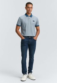 Ljusblå pikétröja med mörkblå krage, kombinerad med mörkblå jeans och vita sneakers. Modellen står med händerna i fickorna.