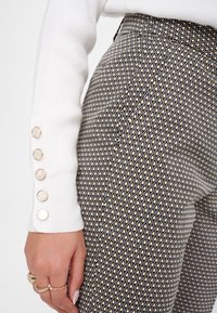 Pantalons à motif noir et or avec une taille haute, présentant un design texturé et une manche côtelée blanche avec des boutons ronds en or.